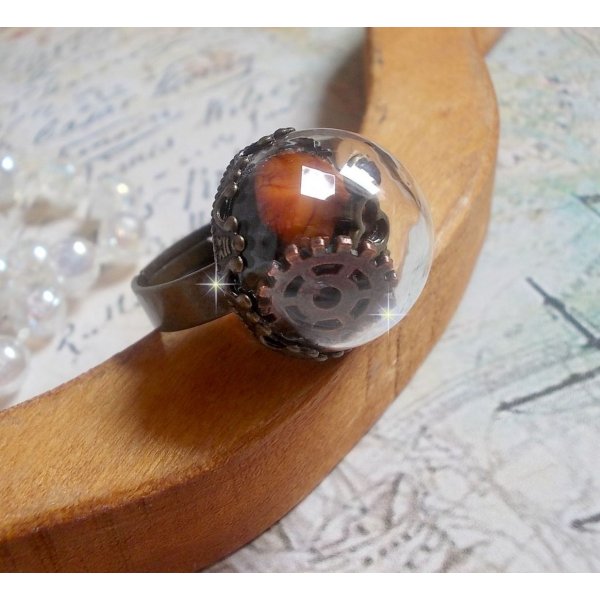Bague Majest&eacute; Steampunk aux rouages m&eacute;caniques &agrave; laquelle s&rsquo;ajoute une cl&eacute; et un strass de couleur Siam et un strass en verre Dor&eacute;. Le tout est mis sous un globe en verre sur une bague r&eacute;glable laiton