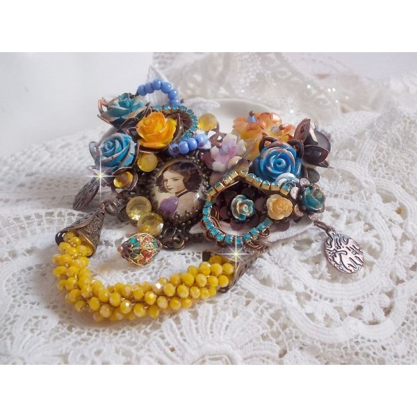 Broche Eclats P&eacute;tillants d&rsquo;inspiration Baroque, orn&eacute;e de fleurs en r&eacute;sine autour d&rsquo;un cabochon strass&eacute;s et perl&eacute;s sur estampes, cristaux, breloques, perle cloisonn&eacute;e, perles jaunes en verre et roses. 