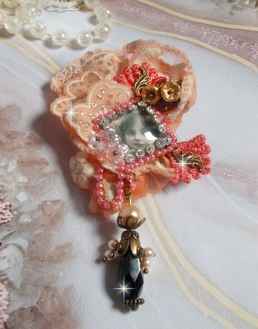 Rustieke rozenbroche gemaakt met een cabochon die een lachend meisje voorstelt, oranje strass- en rozenkant, kristallen, glazen kralen en diverse accessoires.