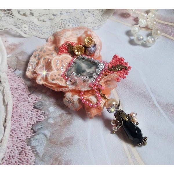 Rustieke rozenbroche gemaakt met een cabochon die een lachend meisje voorstelt, oranje strass- en rozenkant, kristallen, glazen kralen en diverse accessoires.